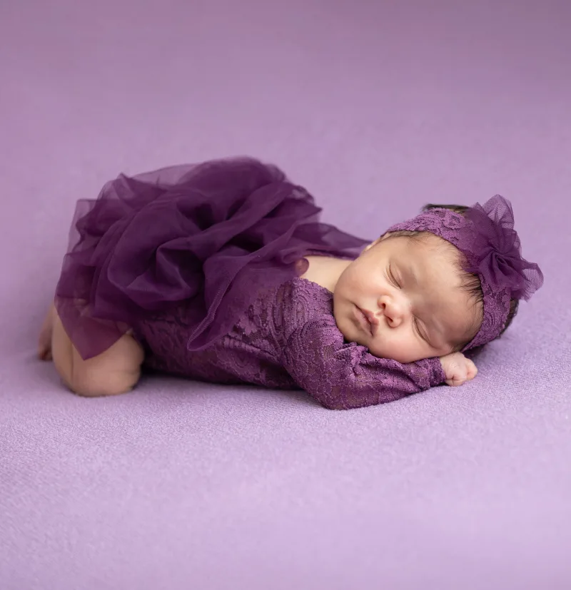 Newborn in a plum lace romper on a lilac backdrop