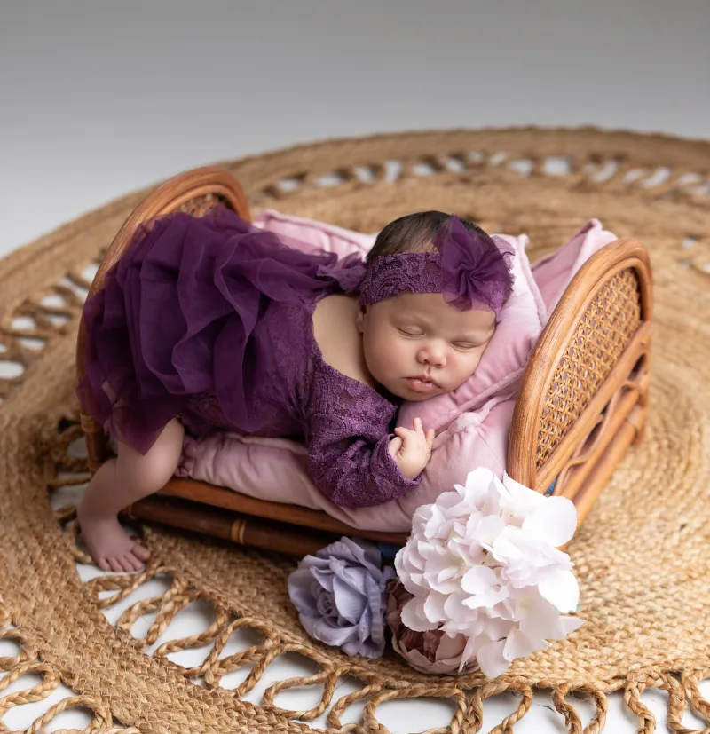 Newborn in a plum lace gown resting on a rattan chair with florals
